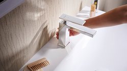 Eine Hand dreht den Wasserhahn in einem modernen Badezimmer, während ein sanftes Licht auf die glänzende Oberfläche fällt.